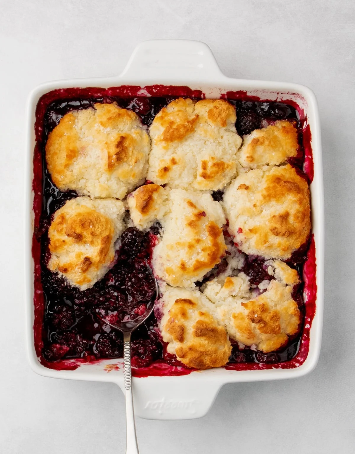 overhead view of baked blackberry cobbler in a square white baking dish with a spoon in the berry filling