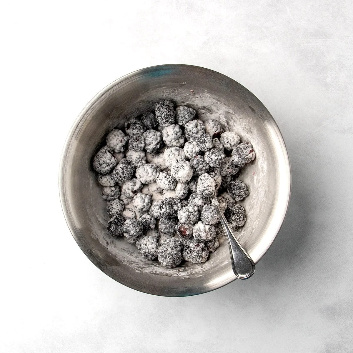 blackberries coated in a white powder mixture in a metal mixing bowl with a spoon