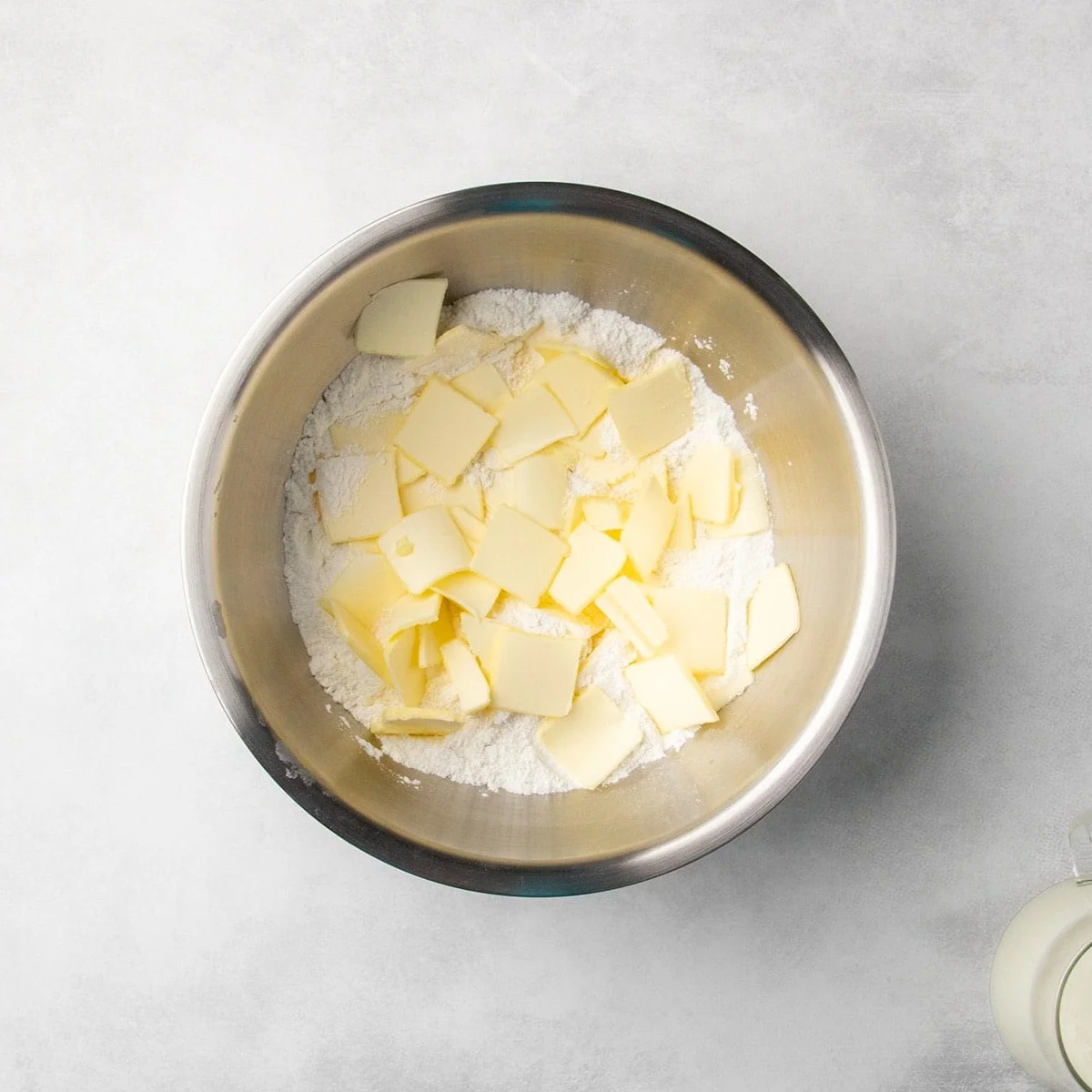 metal mixing bowl with white dry ingredients topped with small, mostly square pieces of sliced butter