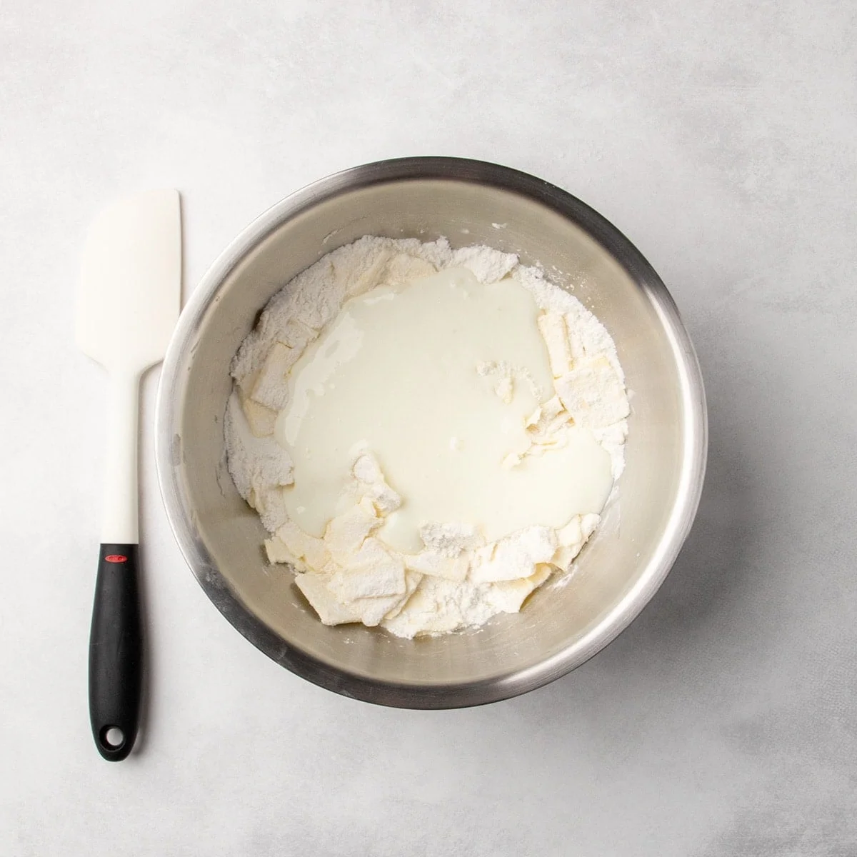 metal bowl with dry ingredients, chunks of butter, and white liquid buttermilk added, spatula placed beside the bowl