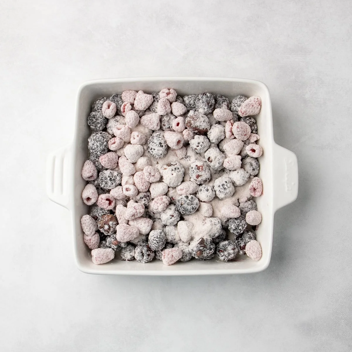 blackberries and raspberries coated in white powder spread in an even layer in a square white baking dish