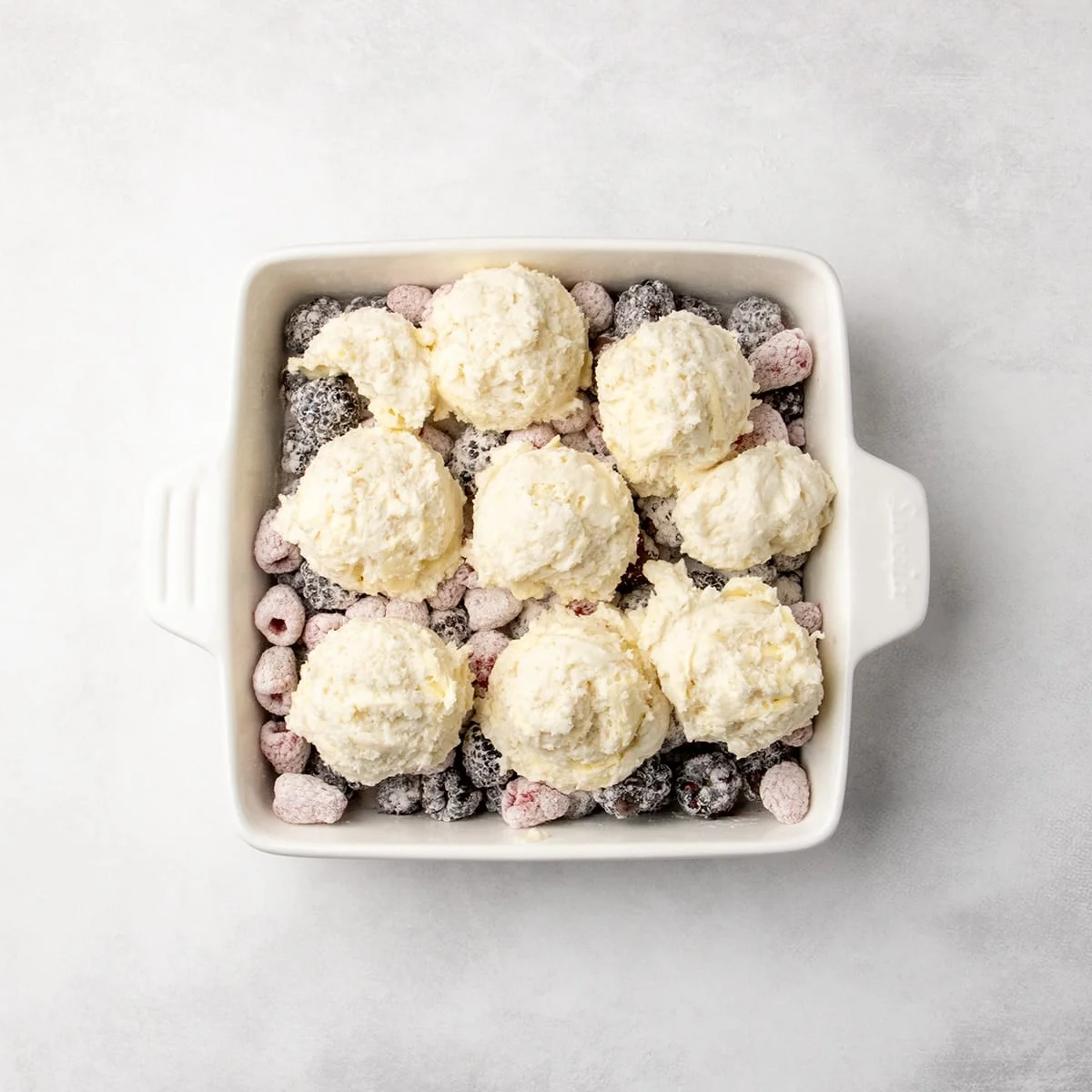 scooped mounds of pale raw dough mixture placed on top of the berry and flour mixture in a white baking dish
