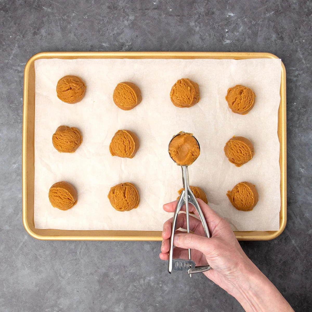 11 brown scoops of peanut butter cookie dough on gold baking sheet lined with white paper, with hand holding metal cookie scoop with cookie dough in the bowl of the scoop