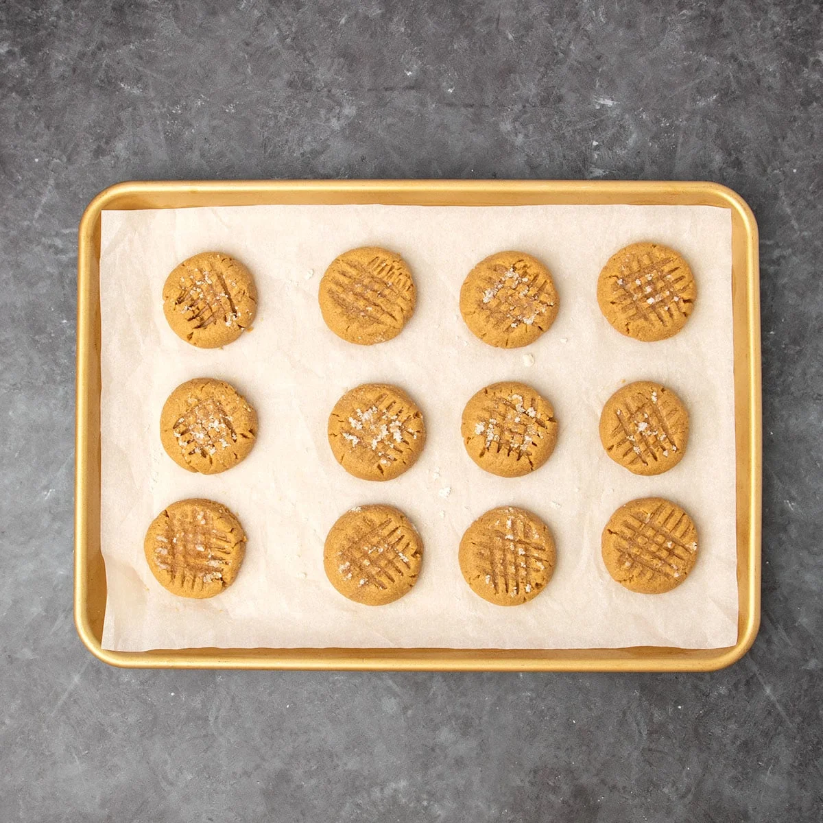 12 freshly baked round, light brown flourless peanut butter cookies with crosshatch pattern on top of each on white paper lined gold baking sheet