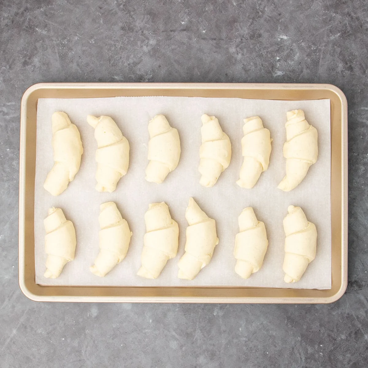 12 small raw crescent shaped rolls of raw dough in 2 rows on white paper on gold colored rectangular jelly roll pan