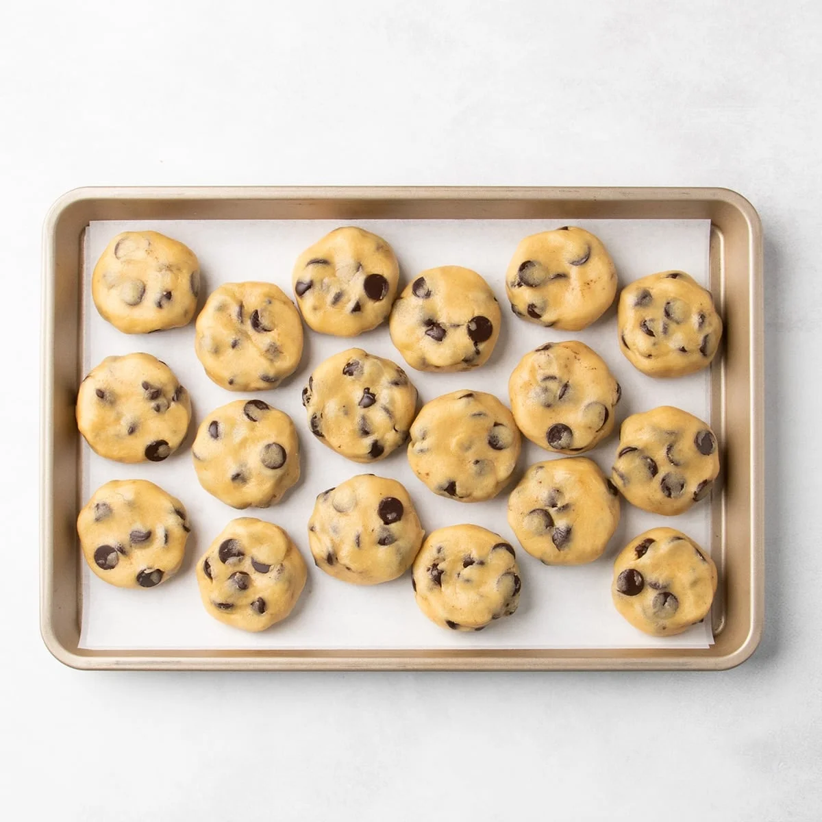 18 flattened raw chocolate chip cookie dough disks arranged on a parchment-lined baking sheet.