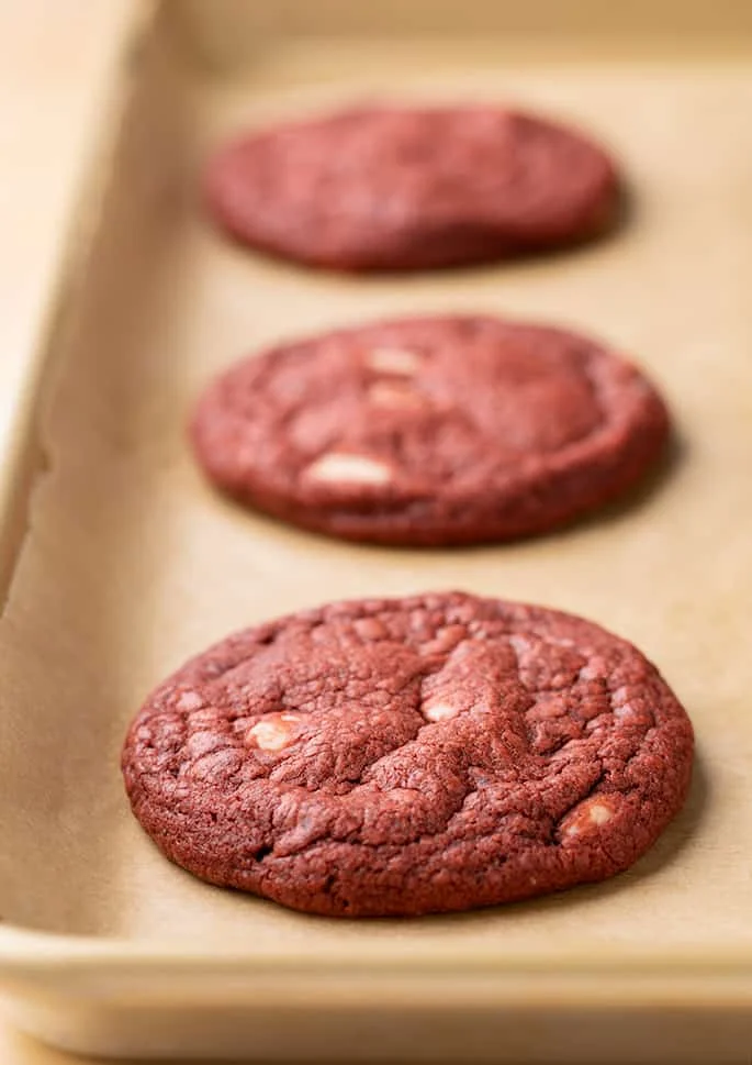 3 baked red round drop cookies with white chocolate chips on brown paper