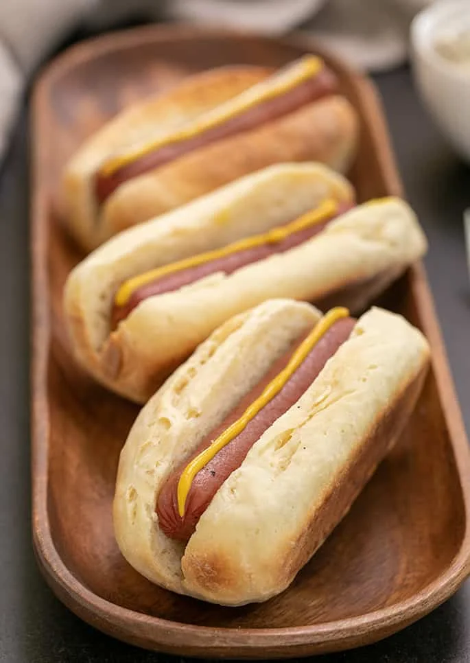 3 hot dogs in buns with mustard on long rectangular wooden tray.