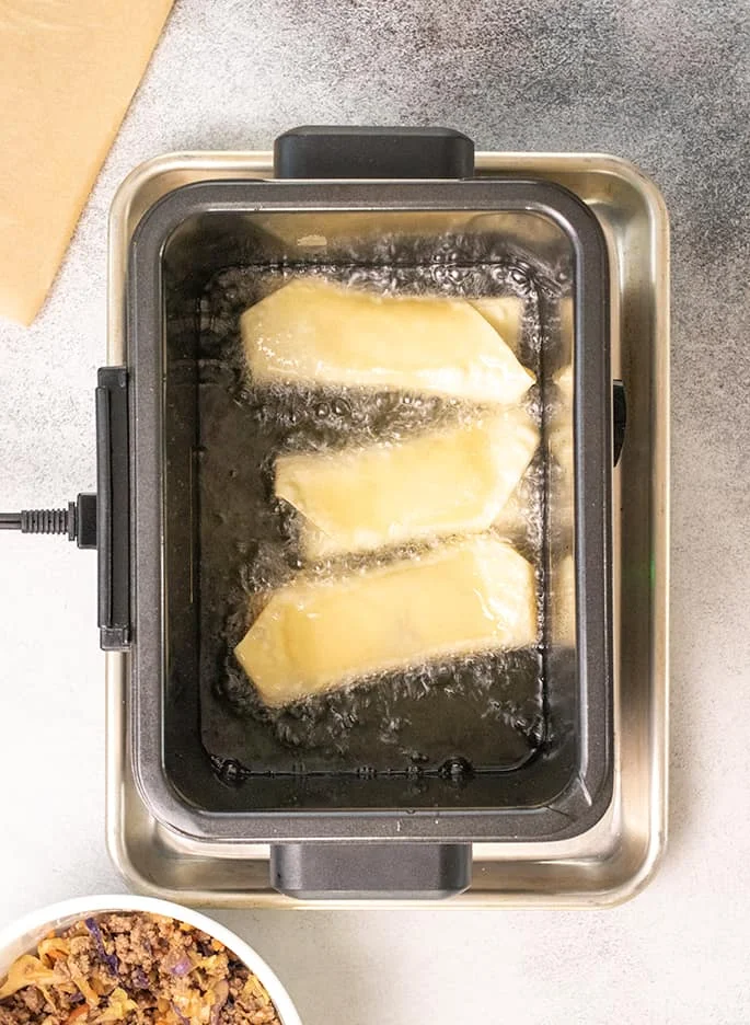 3 raw egg rolls beginning to fry in hot bubbling oil in miniature electric fryer