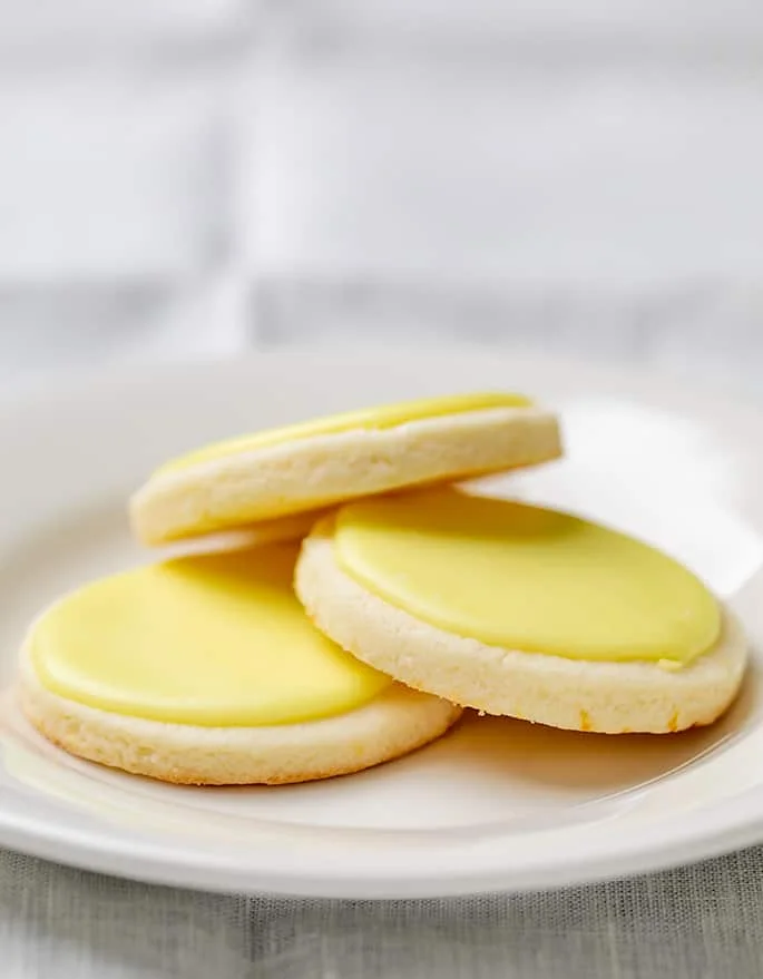 3 round pale cookies with yellow flecks and yellow icing on top on a small white plate on tan cloth