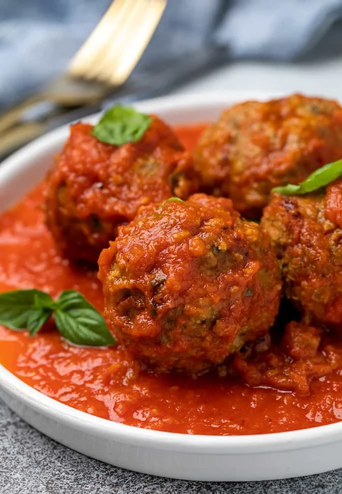 4 cooked meatballs with red sauce on round white plate with fresh basil leaves and a fork and blue cloth in background