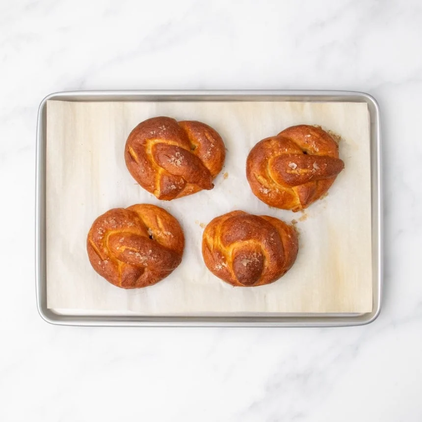 4 fully baked, golden brown gluten free soft pretzels with flaky sea salt on top, all on white paper lining on metal rectangular rimmed baking sheet