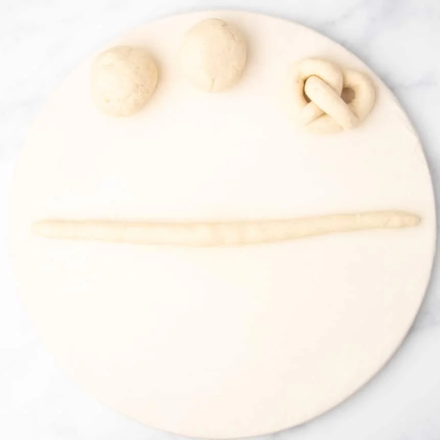 4 pieces of raw bread dough, 2 shaped into loose rounds, one in a pretzel shape, and one rolled out into a 12-inch long rope