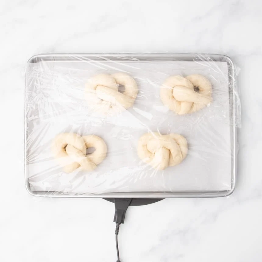 4 raw soft pretzel shapes evenly spaced on baking tray, with tray covered in plastic wrap, set to rise