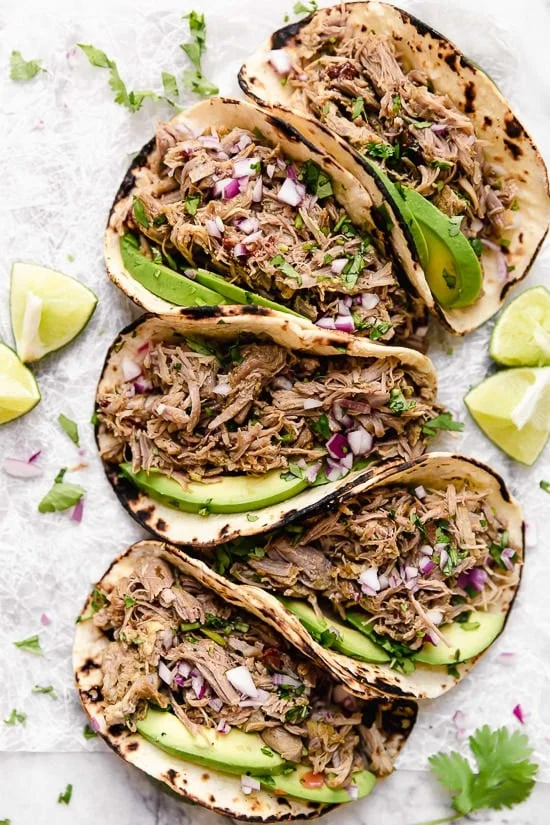5 pork carnitas tacos topped with slices of avocado and red onion.