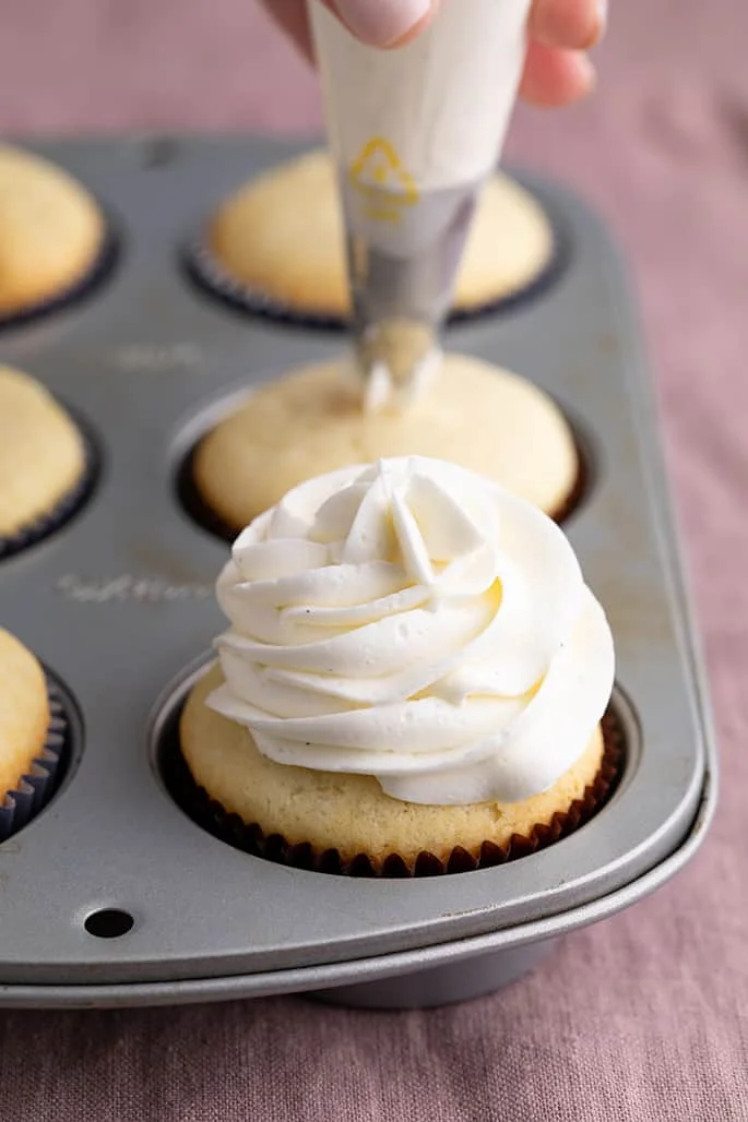6 baked vanilla cupcakes in a muffin tin one with frosting swirl and hand holding piping bag on top of second cupcake in background