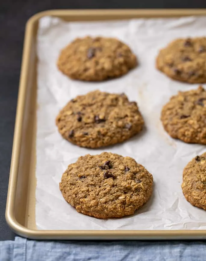 6 brown round oatmeal cookies on white parchment paper on a gold rimmed baking sheet