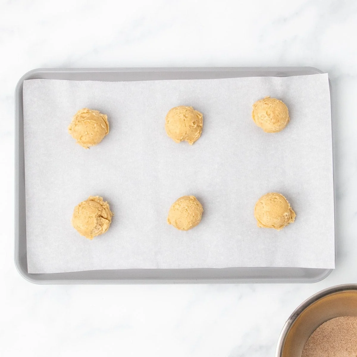 6 scooped portions of raw snickerdoodle cookie dough evenly spaced on white paper lining gray baking sheet on marble counter.