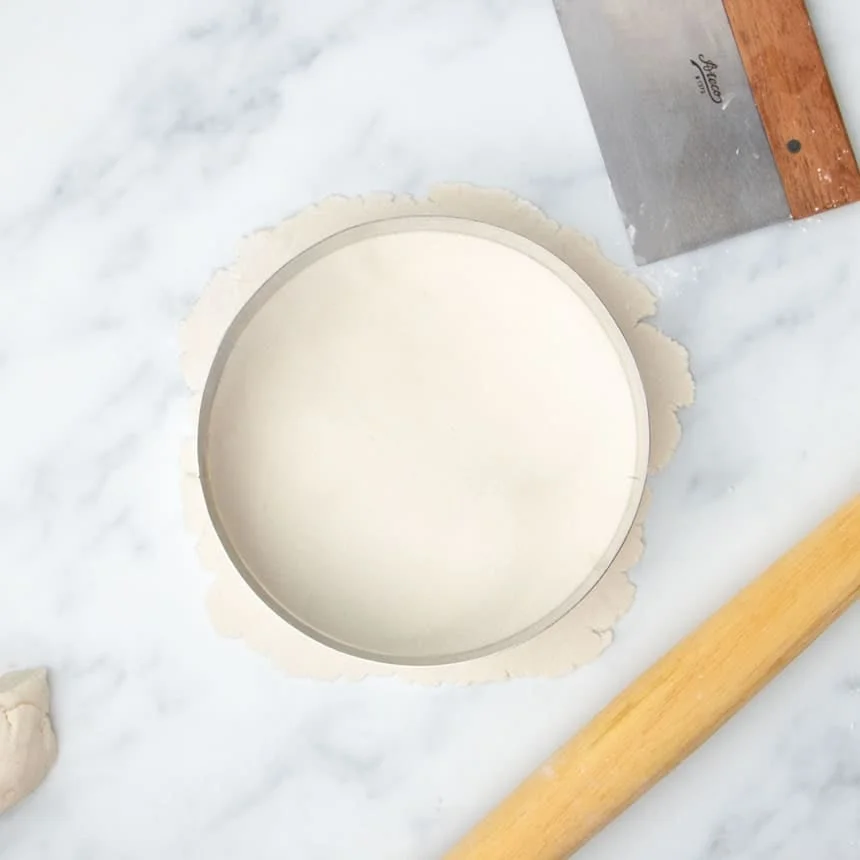 8-inch round metal cake cutter on top of rolled out piece of raw tortilla dough with bench scraper and blond wood tapered rolling pin