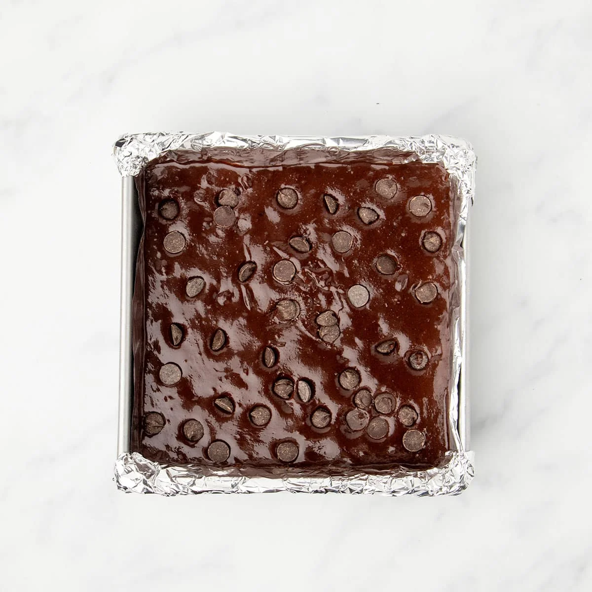 8-inch square metal baking pan lined with foil with dark brown raw brownie mixture with chocolate chips on top.