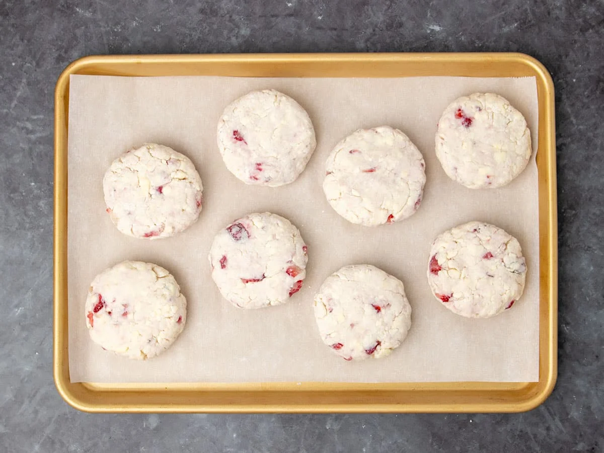 8 raw disks of cream colored biscuit dough with bright red specks of chopped strawberries on white paper on gold baking tray