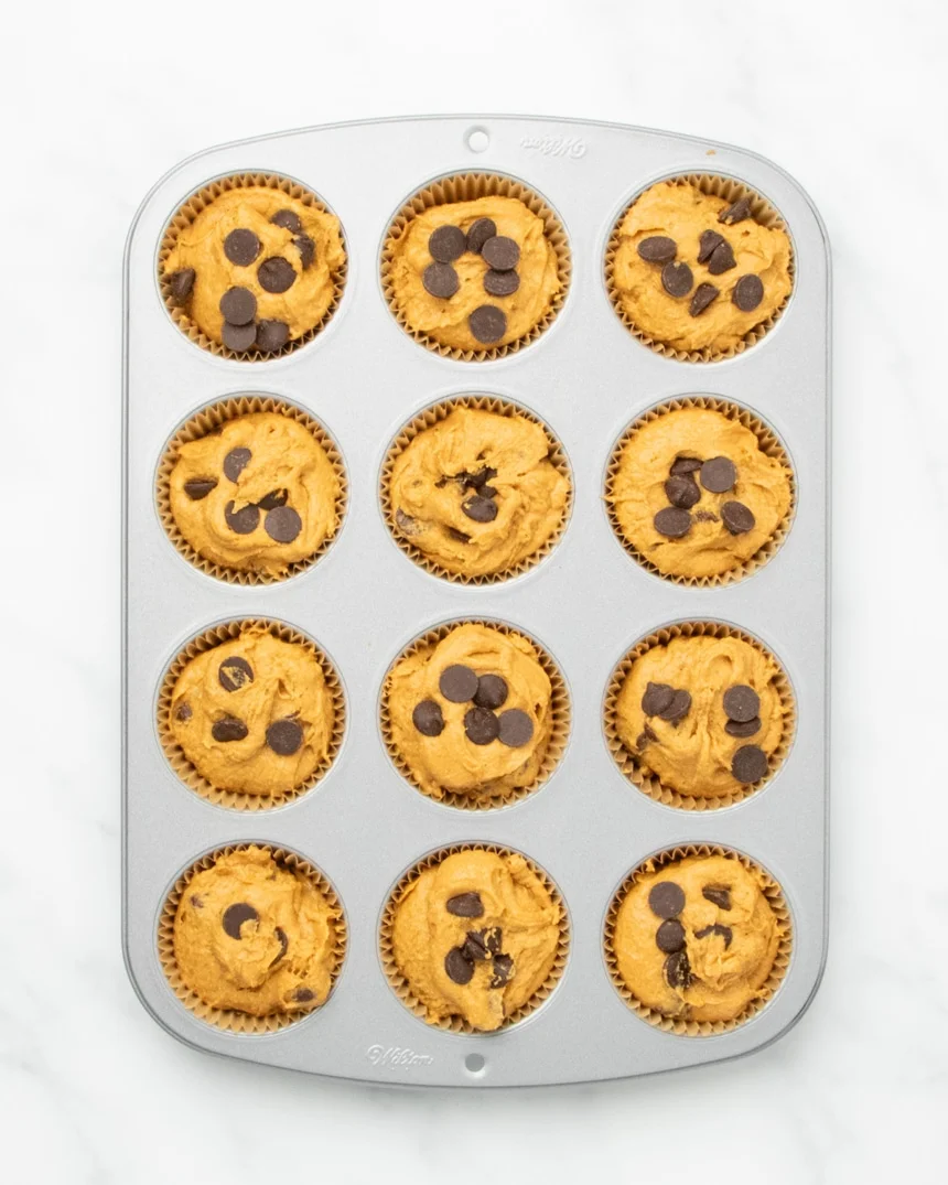 A 12-cup muffin tin with paper liners holds scoops of raw light orange muffin batter topped with dark brown chips.
