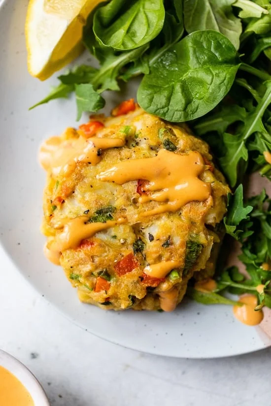 A baked lump crab cake drizzled with red pepper sauce next to a green salad on a plate.
