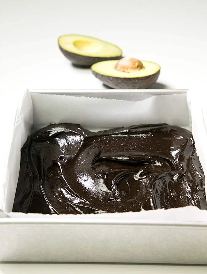 A baking dish of avocado brownie batter and an avocado on white background 