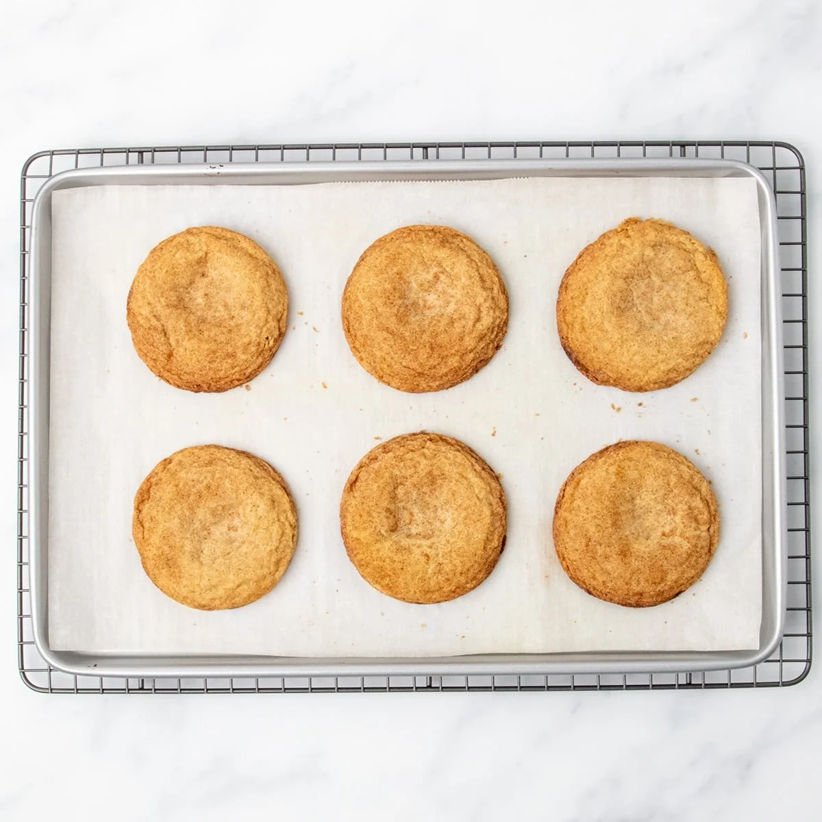 A baking sheet with 6 baked brown round snickerdoodle cookies, cooling on wire rack.