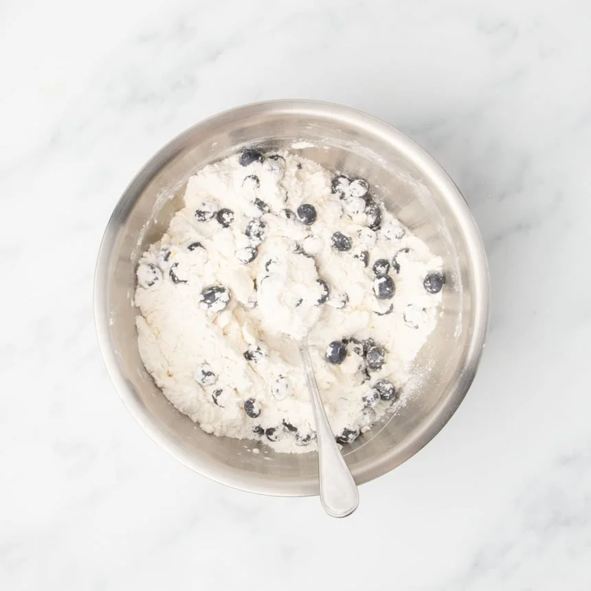 A bowl of dry ingredients and blueberries mixed with a spoon.