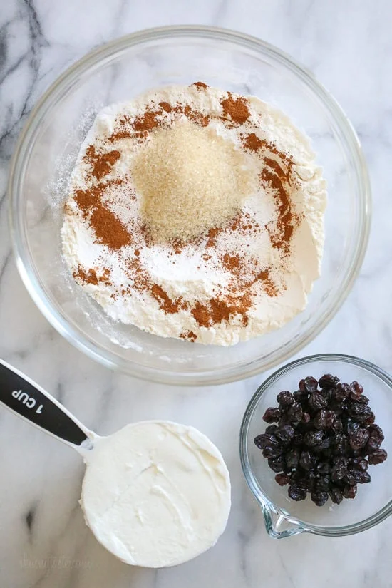 A bowl of dry ingredients next to a measuring cup of Greek yogurt an a bowl of raisins.