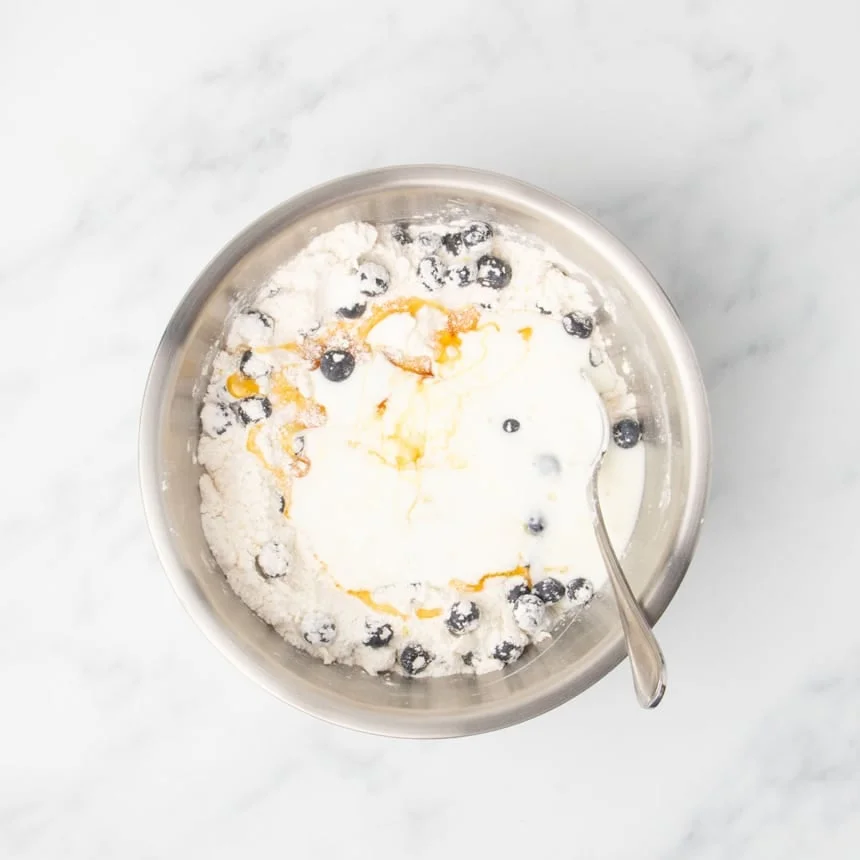 A bowl showing milk, honey, and vanilla poured over the flour, butter, and blueberry mixture.
