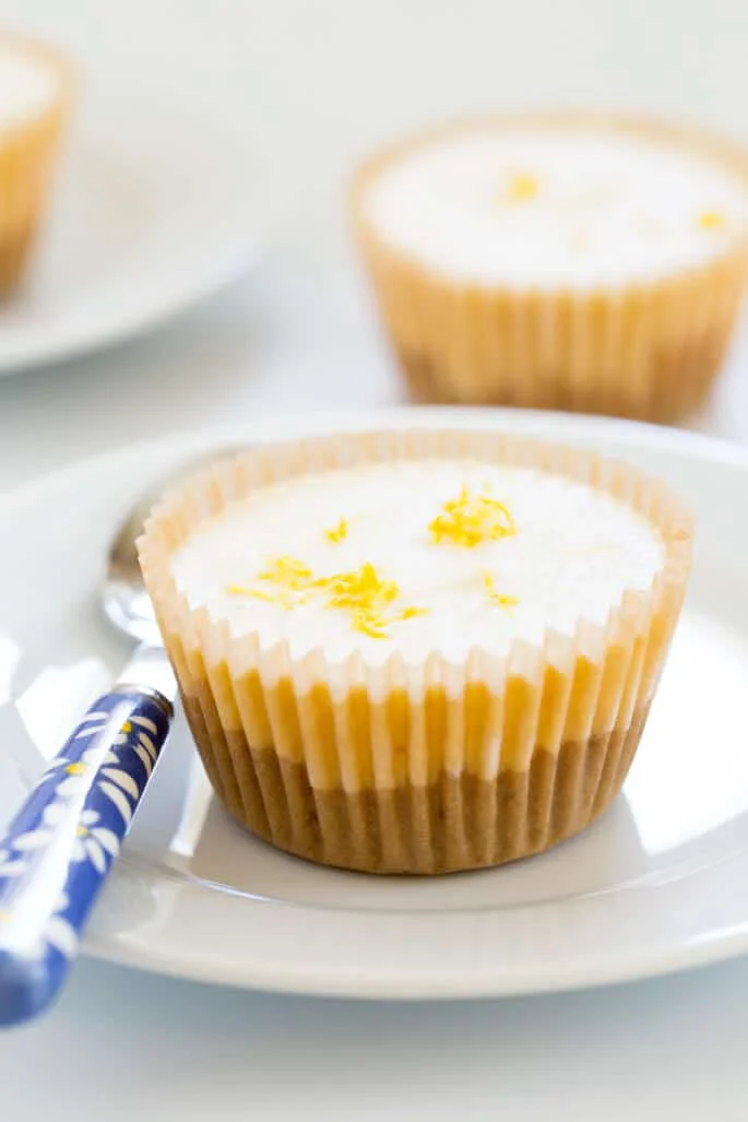 A close up of a lemon cheesecake in a small circular shape on a white plate with a blue spoon