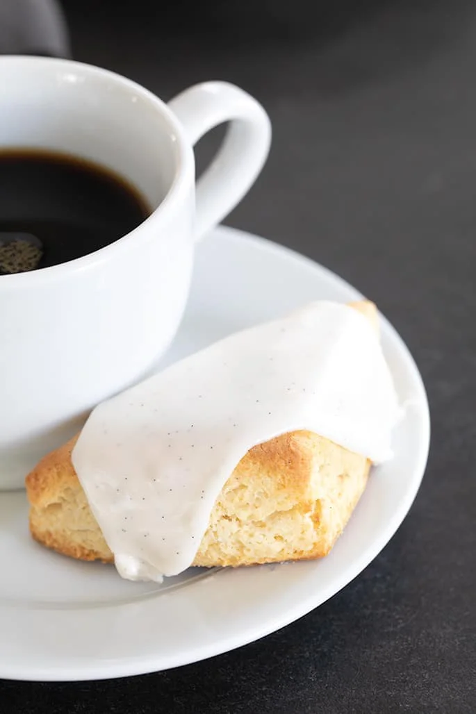 A close up of a vanilla scone with white icing and coffee on a small plate