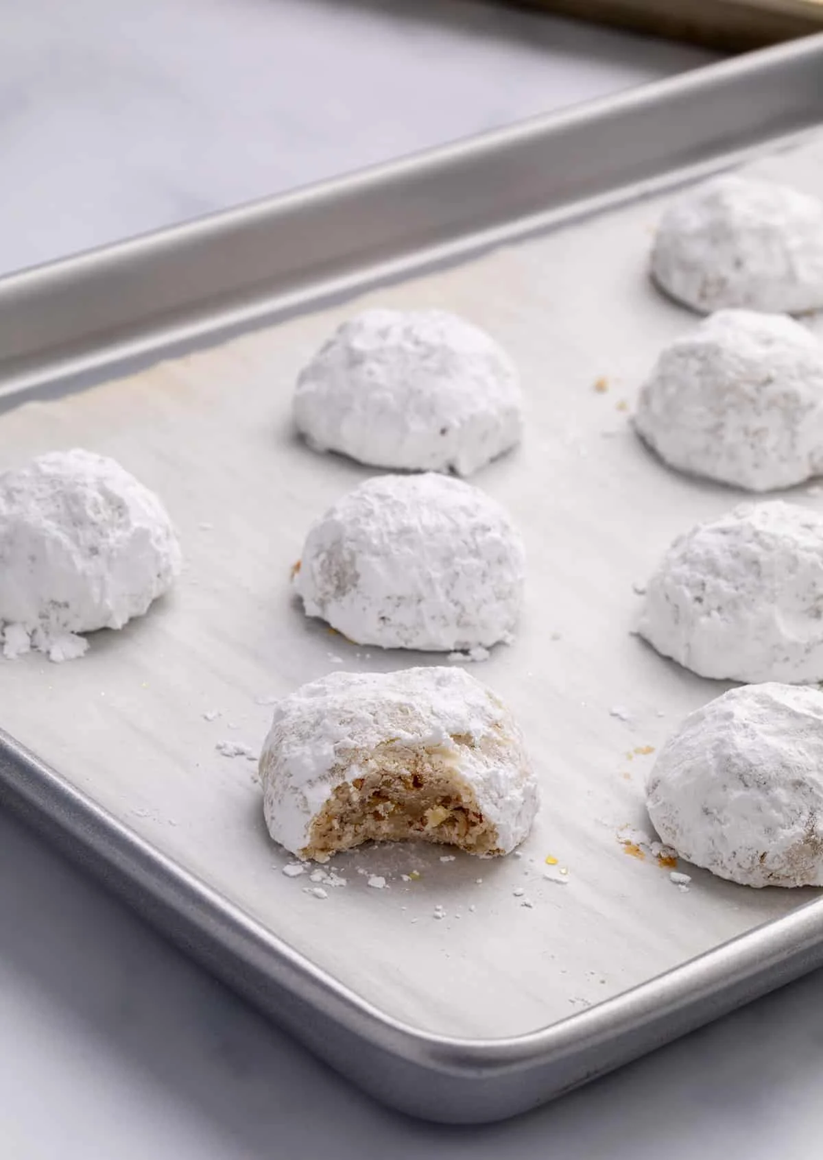 A close view of powdered-sugar-coated cookies on a parchment-lined baking sheet, with one cookie in front showing a bite taken out revealing flecks of chopped pecans inside.