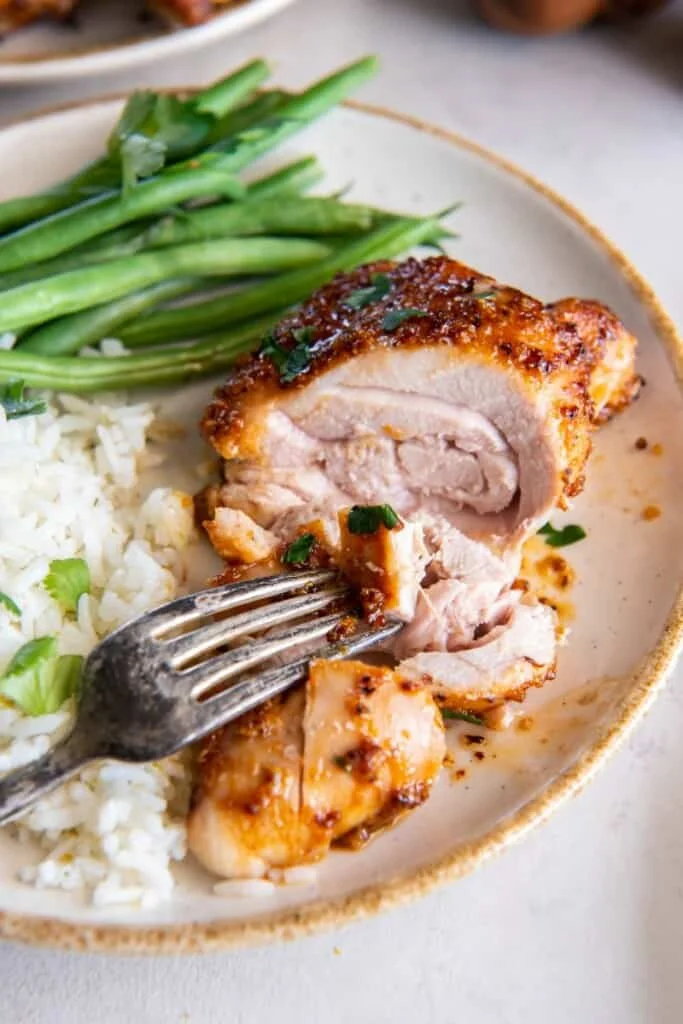 A fork holding a piece of boneless chicken thigh on a white plate with rice and green beans.