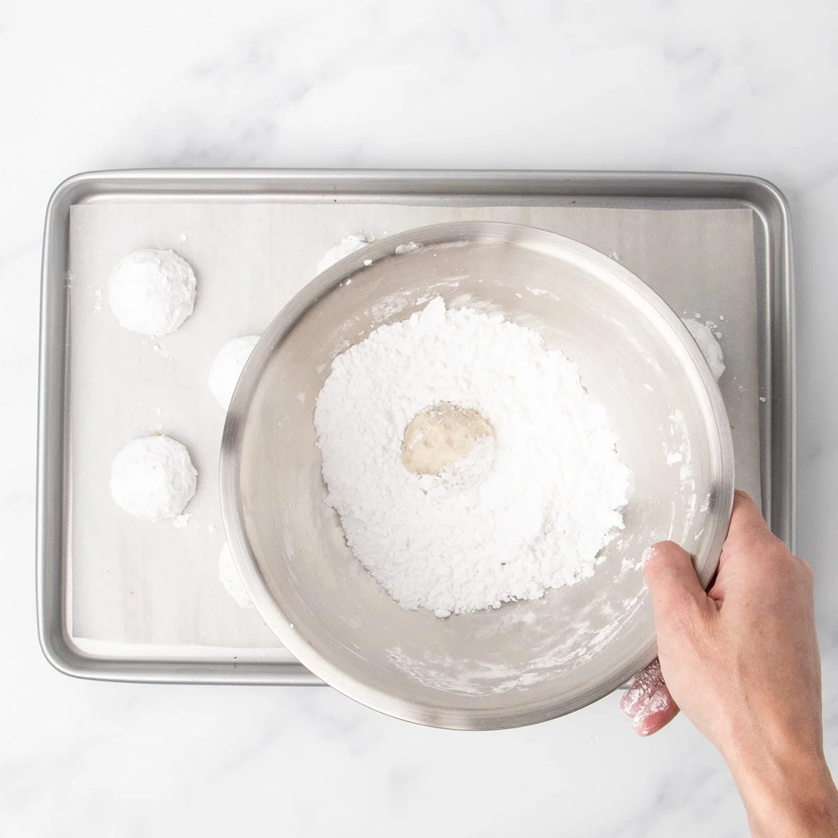 A hand holding a metal bowl filled with powdered sugar, with a baked cookie sitting in with more baked cookies on a parchment lined baking sheet below.