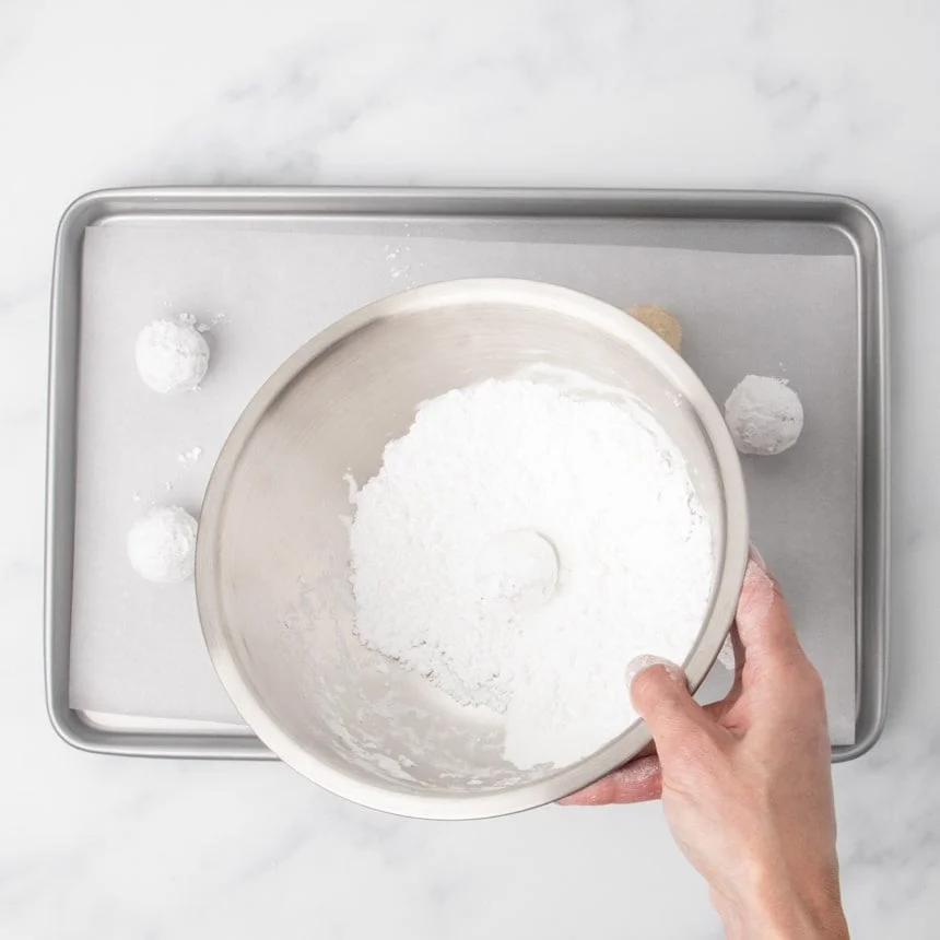 A hand holding a metal bowl partially filled with powdered white sugar, with one dough ball coated in the sugar and other coated dough balls on the baking sheet below.