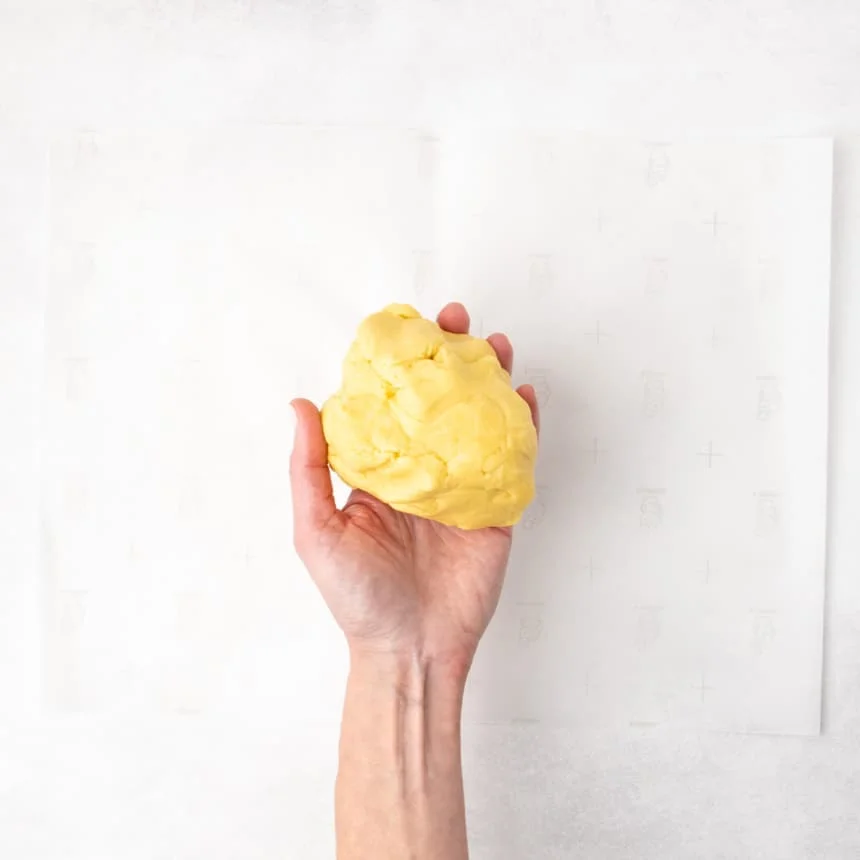 A hand holding a rounded piece of yellow dough over a sheet of parchment paper.