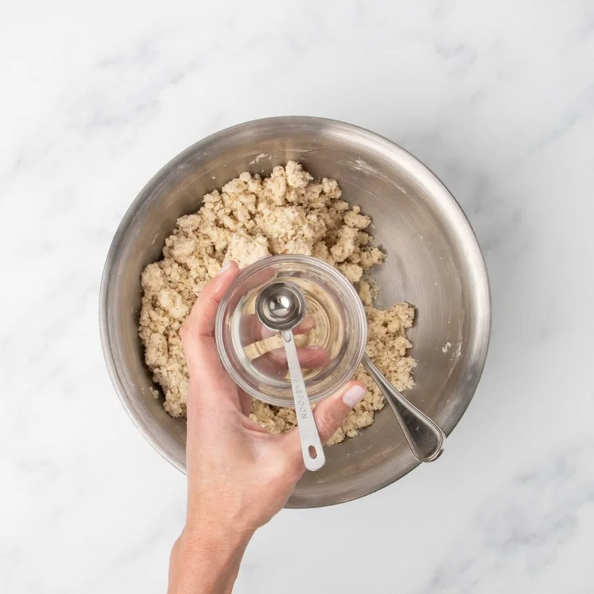 A hand holding a small glass bowl with a metal measuring spoon inside it over a larger bowl of clumps of light tan raw cookie dough.