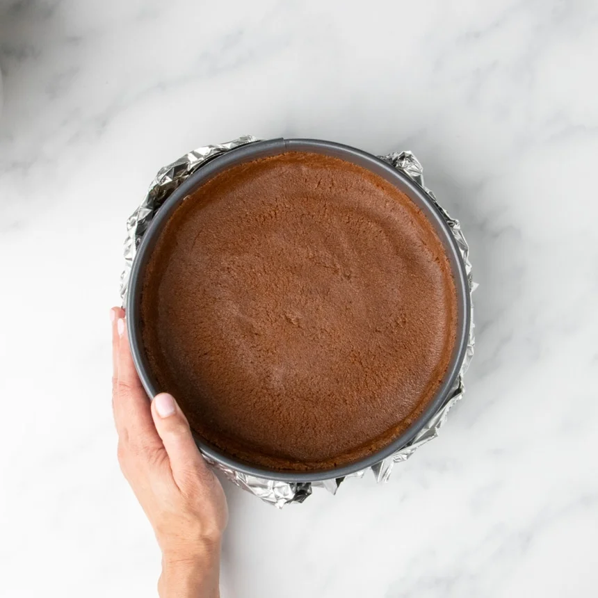 A hand holding a springform pan with a prepared cookie crust, now wrapped in aluminum foil to prepare it for baking.