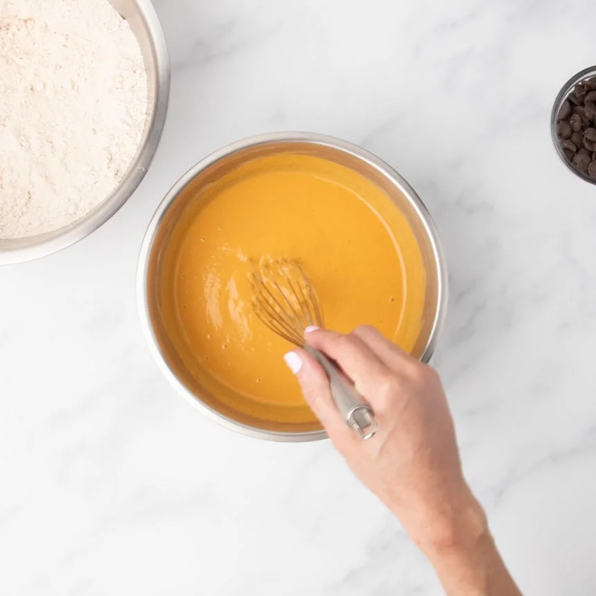 A hand holding a whisk being used to mix the ingredients in the bowl, which are now a smooth orange color