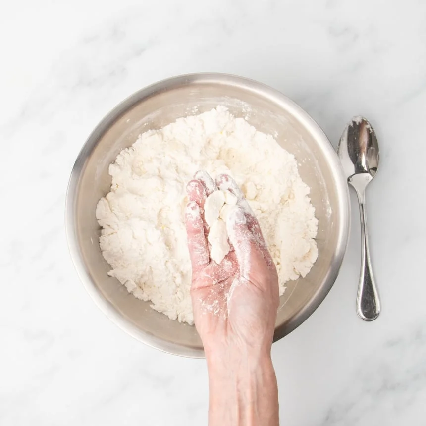 A hand holding clumps of butter and flour mixture, showing how to "cut" the butter into the dough by pressing the clumps flat.