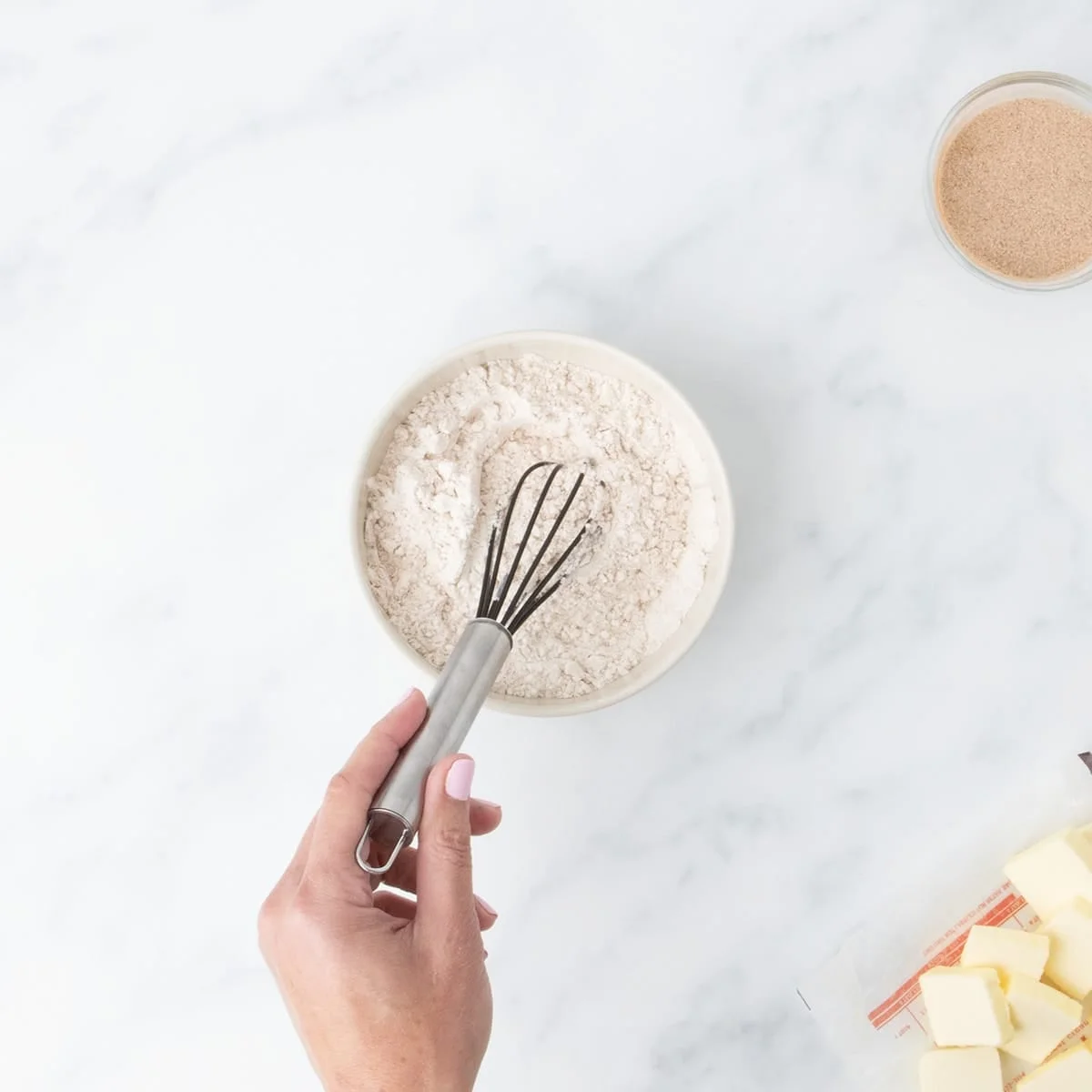 A hand holding small balloon whisk in small bowl of dry white flour mixture on marble surface.