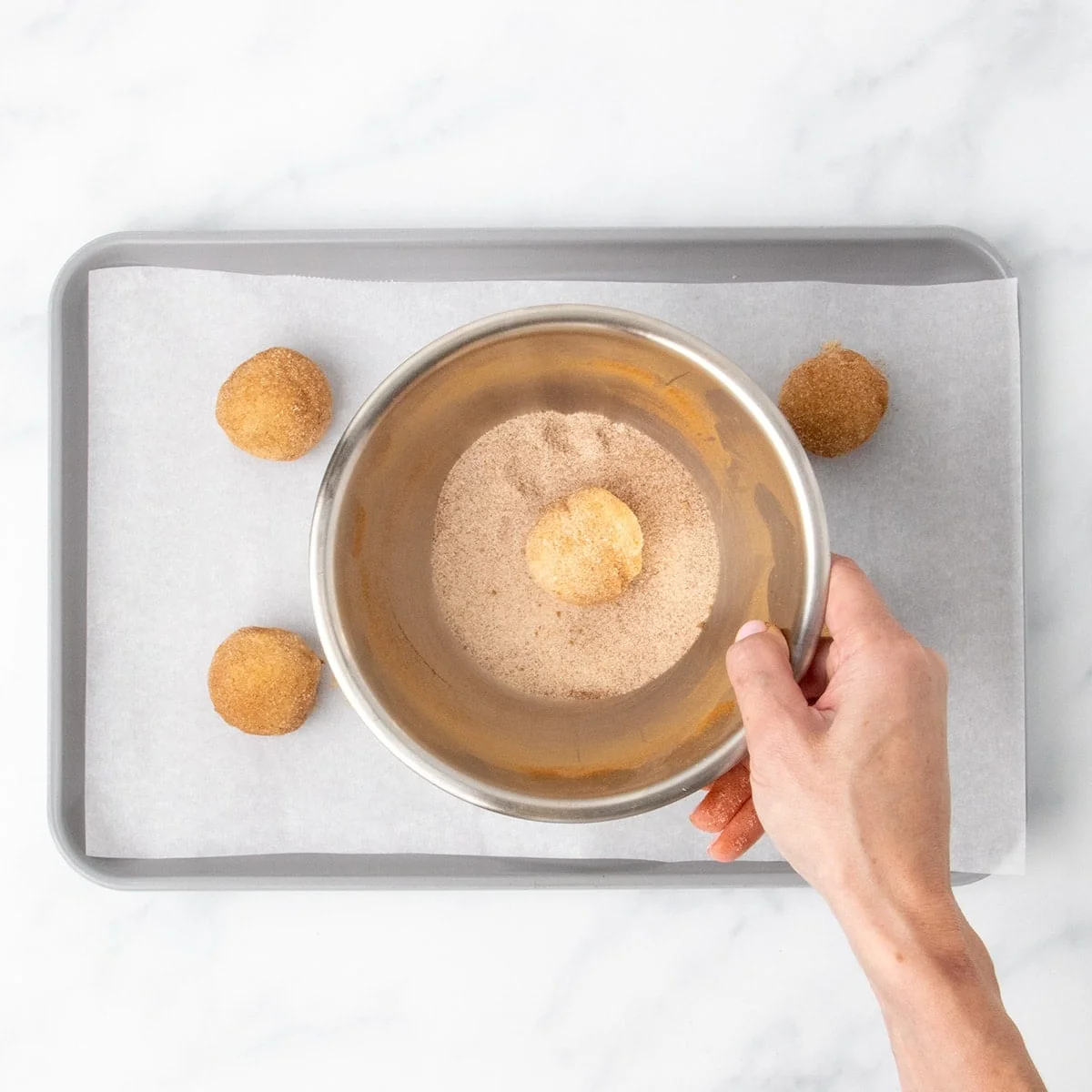 A hand holding small metal bowl with cinnamon sugar mixture, covering ball of cookie dough in mixture.