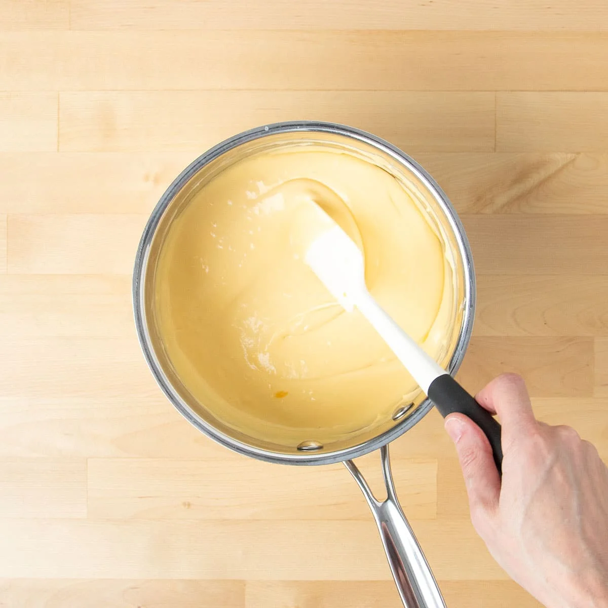 A hand holding white spatula mixing and turning over light yellow cheese sauce in saucepan.