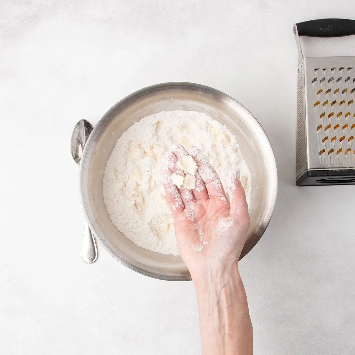 A hand holds flattened shards of butter over a metal mixing bowl filled with white whisked dry ingredients; a box grater sits to the right of the bowl.