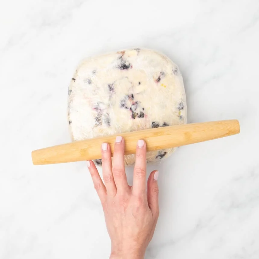 A hand using a rolling pin to gently flatten the dough round while it is covered in plastic wrap.