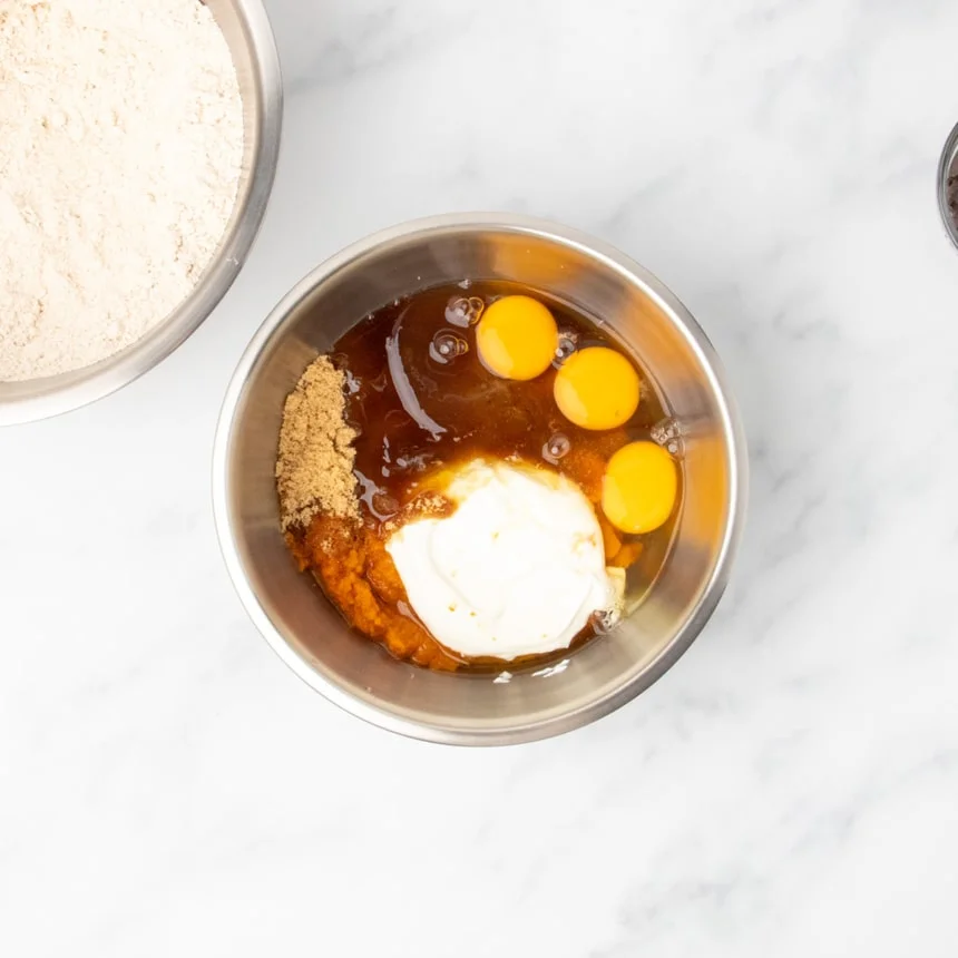 A medium-size round metal mixing bowl contains brown sugar, pumpkin puree, eggs, and a sour cream, unmixed.