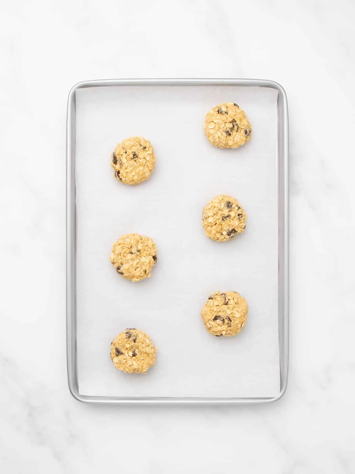 A metal baking sheet lined with white parchment paper, holding six evenly spaced unbaked disk-shaped mounds of cookie dough.
